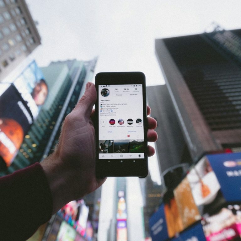 Eine Hand hält ein Smartphone, vor großen Werbetafeln in einer Stadtlandschaft.